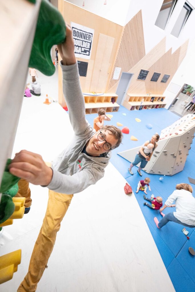 Ein Mann hält sich an einer Kletterwand fest und schaut fröhlich in die Kamera